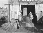 Two Men Working With Bait For Lobstering, On Dock With Barrels In Front Of Building At New Harbor by George French
