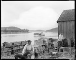 Two Men Work On Traps On Shore, One Sits With Dog, Shack On Right, Traps On Boat Out In Harbor At Bucks Harbor by George French