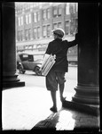 Back To View Of Newsboy, In City, Leaning Against Pillar Of Bld. ,looks Out On Street. by George French