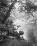 Man Sitting On Stream Bank Holding Fish, Another On Rock Beside Him by French George
