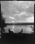 Two People Sitting In Lawn Chairs On Lake Shore At Sunset, Mountains In Distance In Lovell by George French