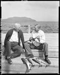 Two Men On Wharf Talking, One With Fishing Rod And Fish He Caught At Moosehead by George French