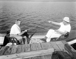 Two Men In A Canoe By Dock Telling Fish Stories by George French