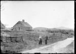 Couple Standing On The Side Of A Rural Road In Front Of Farmhouse With Fenced Fields by George French