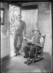 Man Holding A Camera Showing Photographs To A Man Sitting In A Rocking Chair by George French